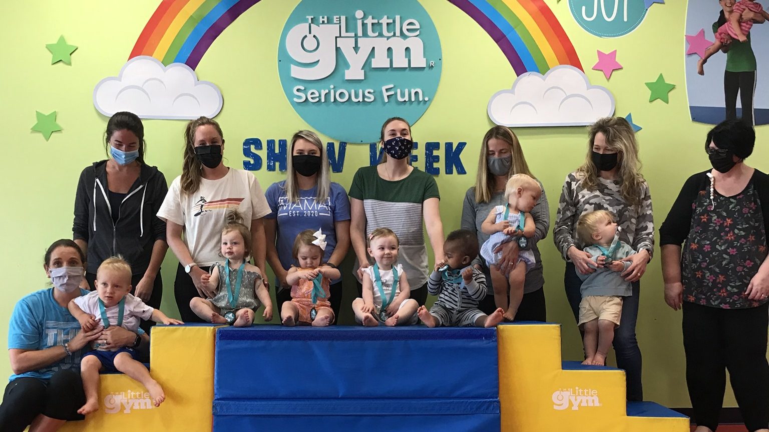 A group of adults and young children wearing masks, posing for a photo at The Little Gym, standing in front of a colorful wall with the gym's logo and rainbow decor.