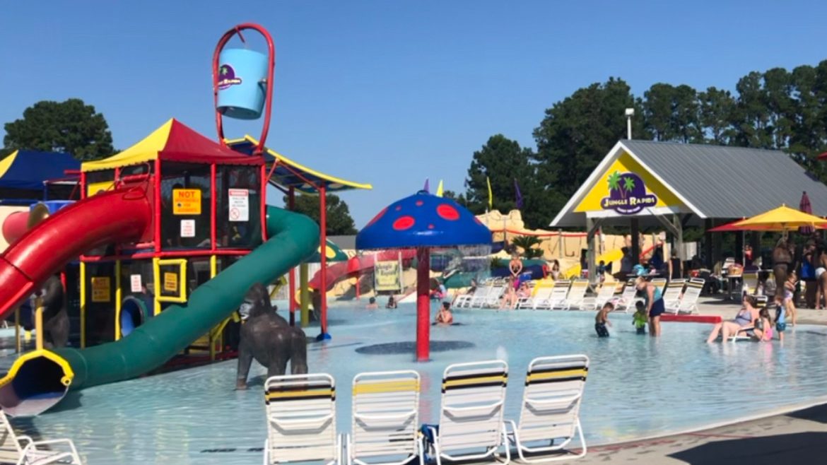 A vibrant water park scene at Jungle Rapids, featuring colorful slides, a splash pad with a large blue mushroom, and families enjoying the water activities on a sunny day.