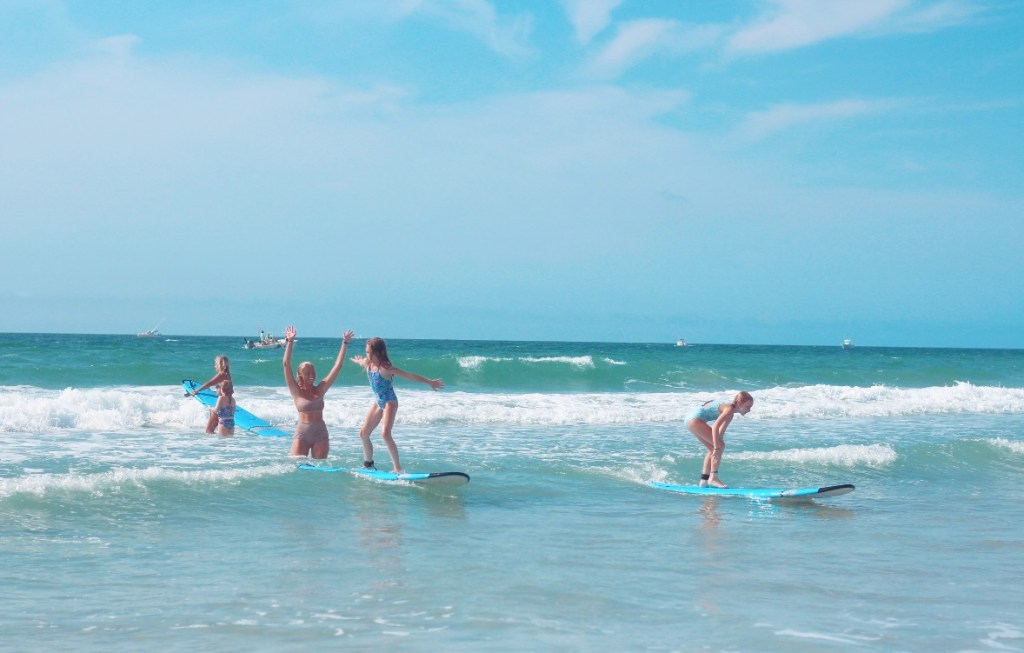 A group of children enjoying a day at the beach, with some surfing on blue boards and others cheering in the water under a clear blue sky.