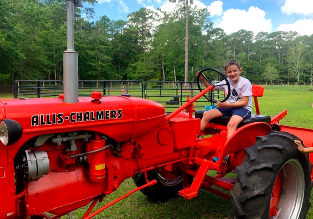 A child sitting on a bright red Allis-Chalmers tractor in a grassy field, surrounded by trees under a blue sky.