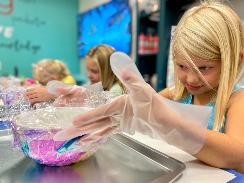 A young girl wearing gloves carefully molds a colorful bowl while working on an art or craft project, with other children engaged in similar activities in the background.