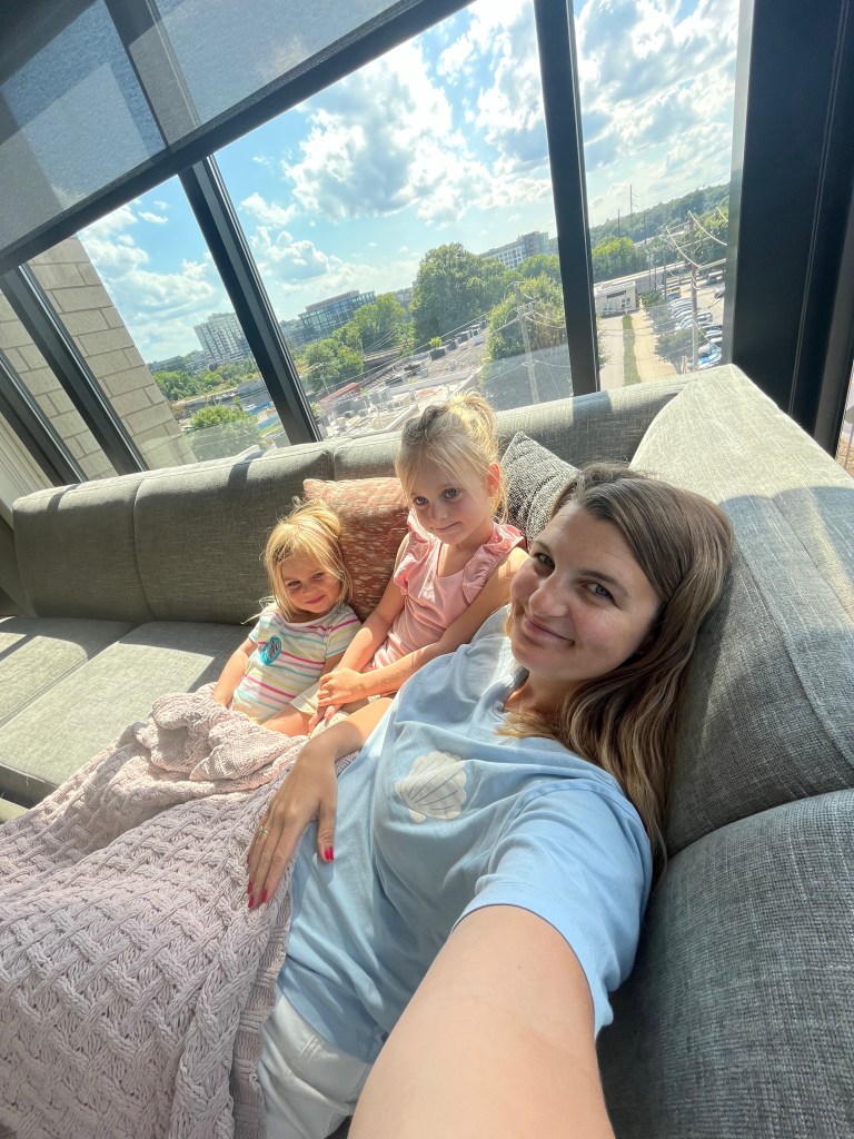 A mother and her two young daughters are sitting on a couch by a large window, enjoying a cozy moment together. The background shows a bright sky and greenery outside.