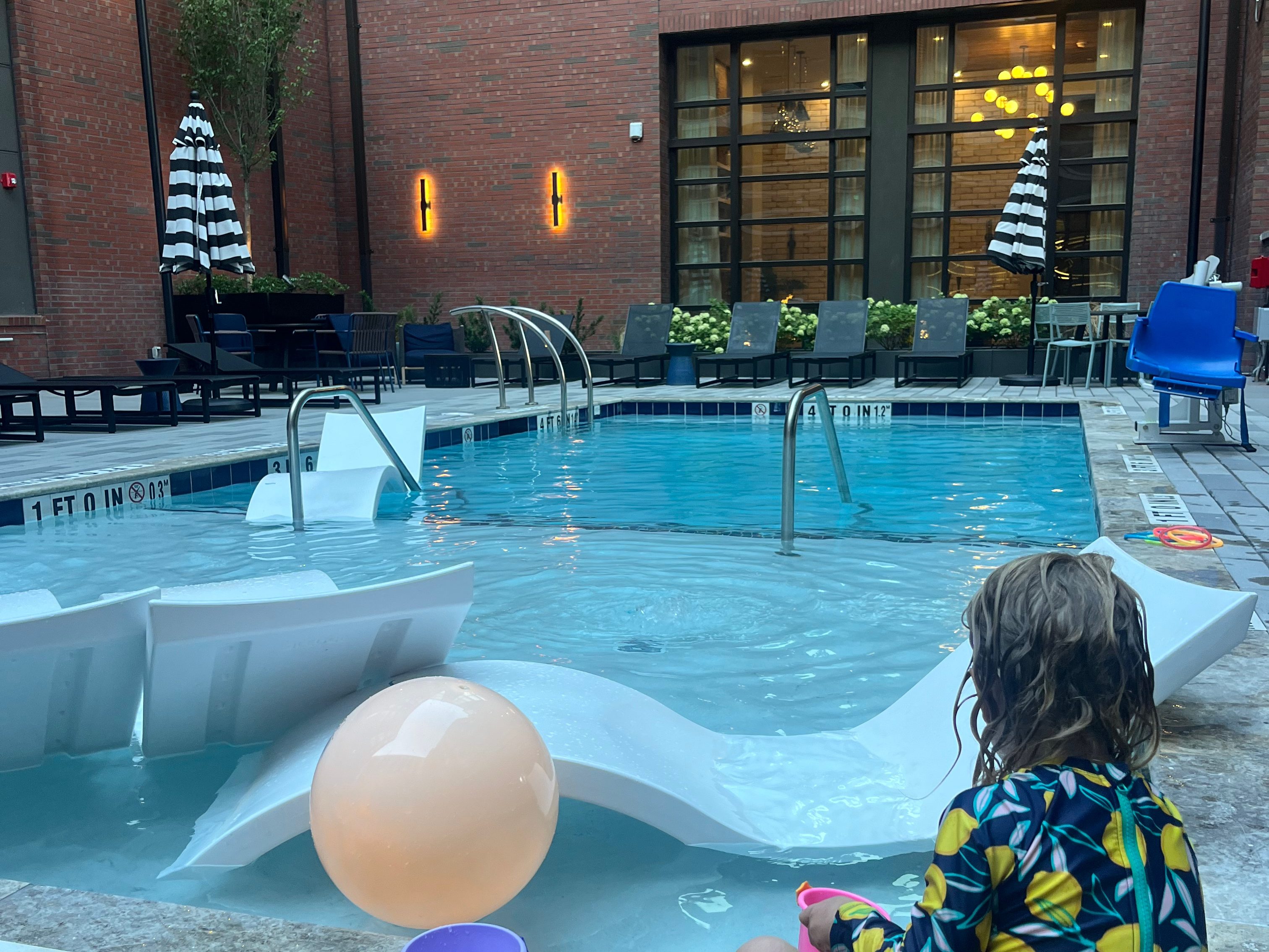 A young child, wearing a colorful swimsuit, sits by a hotel pool, playing with a purple bucket and a pink cup. In the background, lounge chairs and umbrellas are set up around the pool area, showcasing a cozy outdoor space.