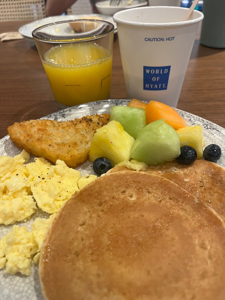 A breakfast plate featuring scrambled eggs, a golden pancake, fruit pieces including cantaloupe and pineapple, and a hash brown. Accompanied by a glass of orange juice and a coffee cup from Hyatt.
