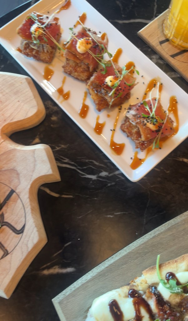 A close-up view of a gourmet dish featuring several pieces of sushi-style food atop a white plate, drizzled with sauce and garnished with green herbs. A wooden serving board is partially visible in the foreground.