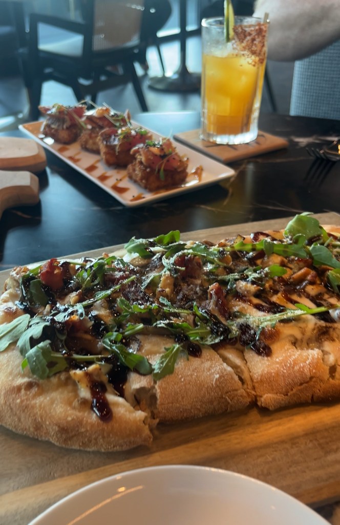 A close-up view of a flatbread topped with greens and drizzled sauce, served on a wooden board, with appetizers and a drink in the background.