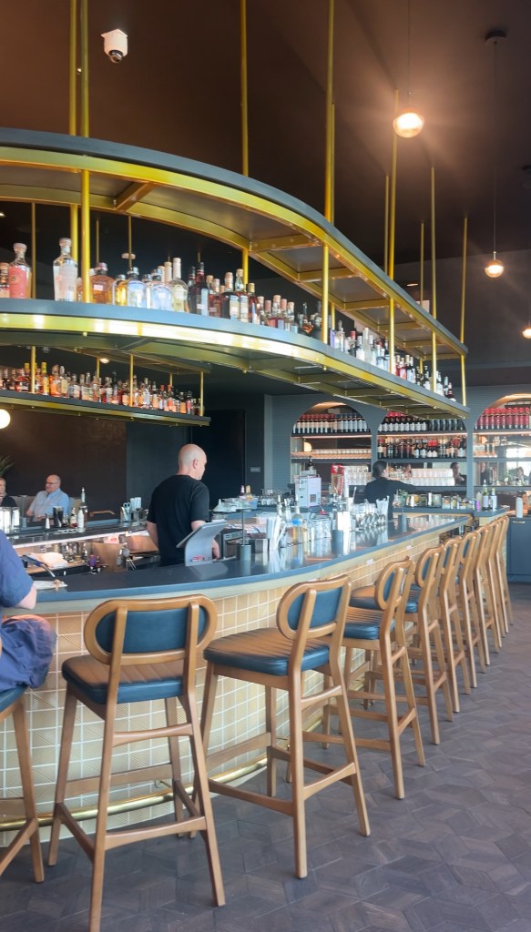 Interior view of a modern bar featuring high stools and a long counter, with shelves displaying various bottles of spirits.