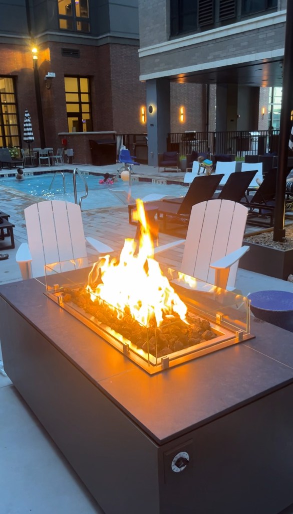 Fire pit with clear glass enclosure in an outdoor pool area, featuring lounge chairs and swimming pool in the background.