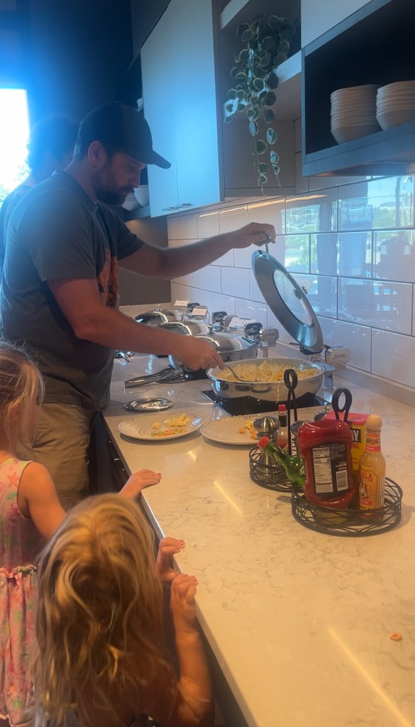 A person cooking in a kitchen while two children watch closely, with plates ready on the counter.