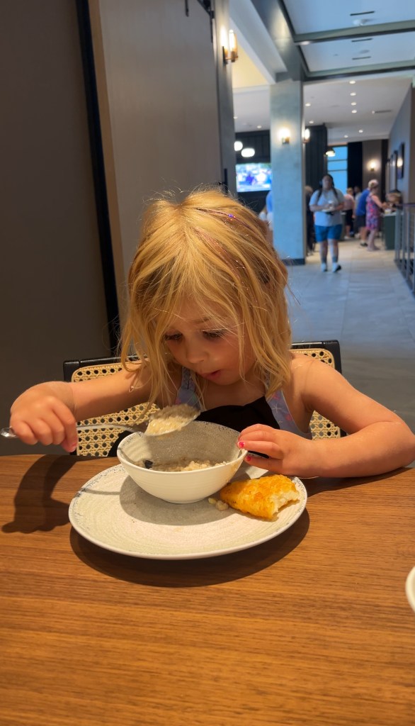 A young child sitting at a wooden table, eating oatmeal from a bowl while holding a spoon, with a croissant on the plate next to her. The setting is a dining area with others in the background.
