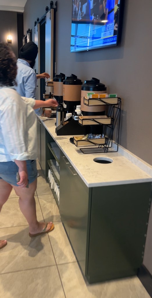 A guest serving themselves coffee at a self-serve station in a modern hotel lobby. There are multiple coffee dispensers and condiments available, with a TV showing news in the background.
