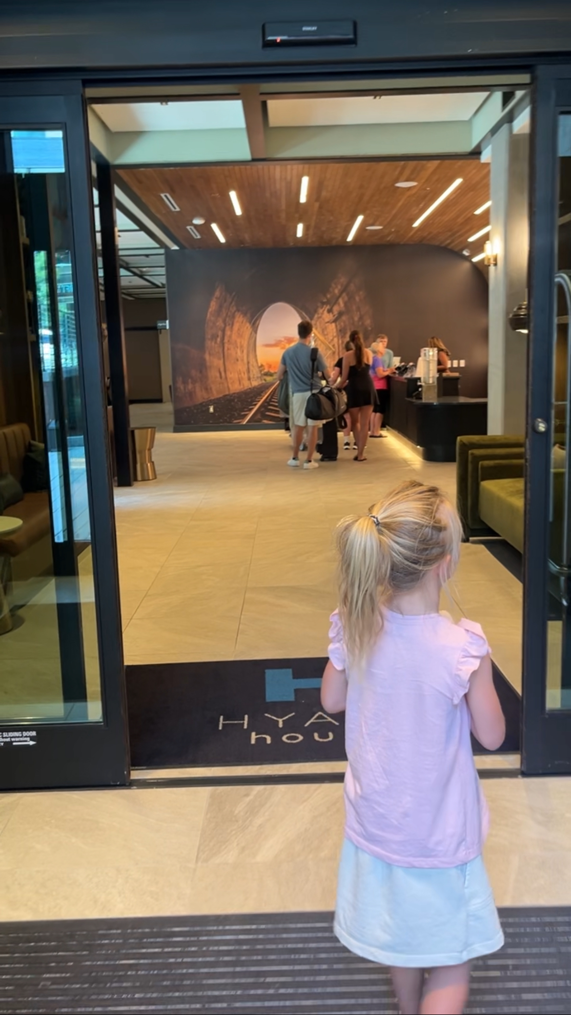 A young child entering the Hyatt House hotel lobby, with guests checking in at the reception desk in the background.