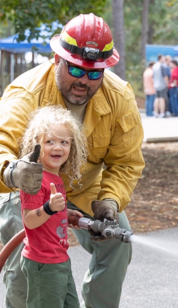 A firefighter and a child giving thumbs up while holding a fire hose at an outdoor event.
