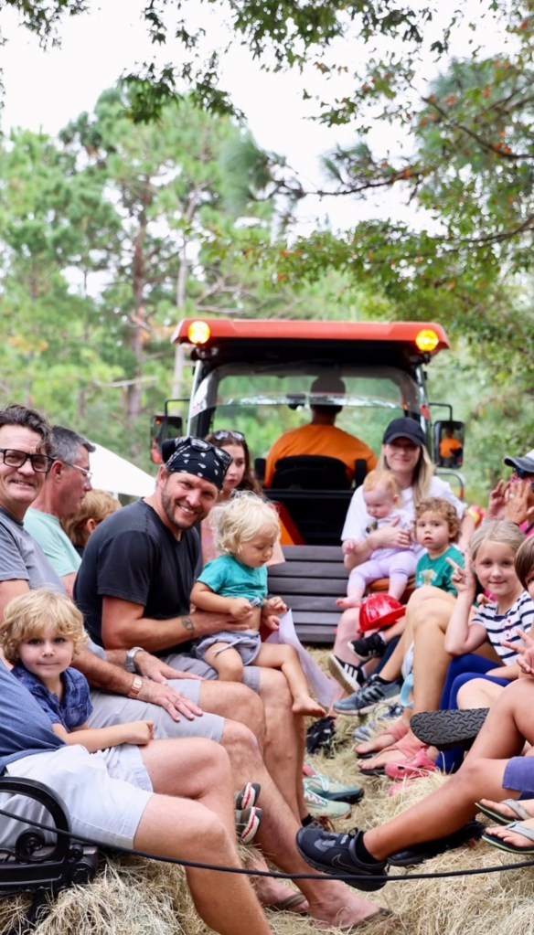 A group of families sitting together on hay bales, enjoying a festival ride in a tractor-drawn wagon. Children and adults are smiling and interacting, surrounded by a natural setting with trees in the background.