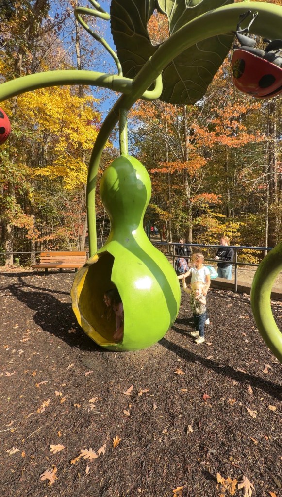 Children playing in a vibrant green playground structure shaped like a pod, surrounded by colorful autumn leaves.