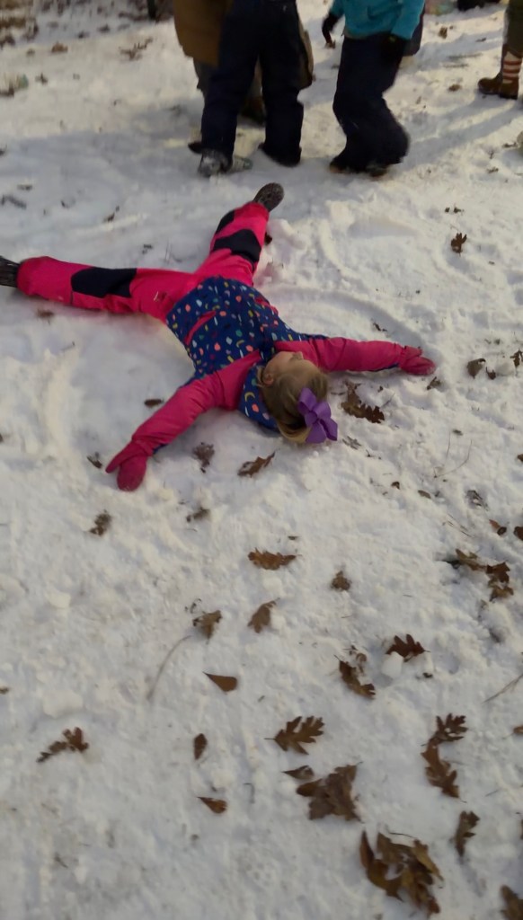 A child wearing a colorful snow suit and a purple bow is lying in freshly made snow, making a snow angel. Surrounding leaves are scattered across the snowy ground.
