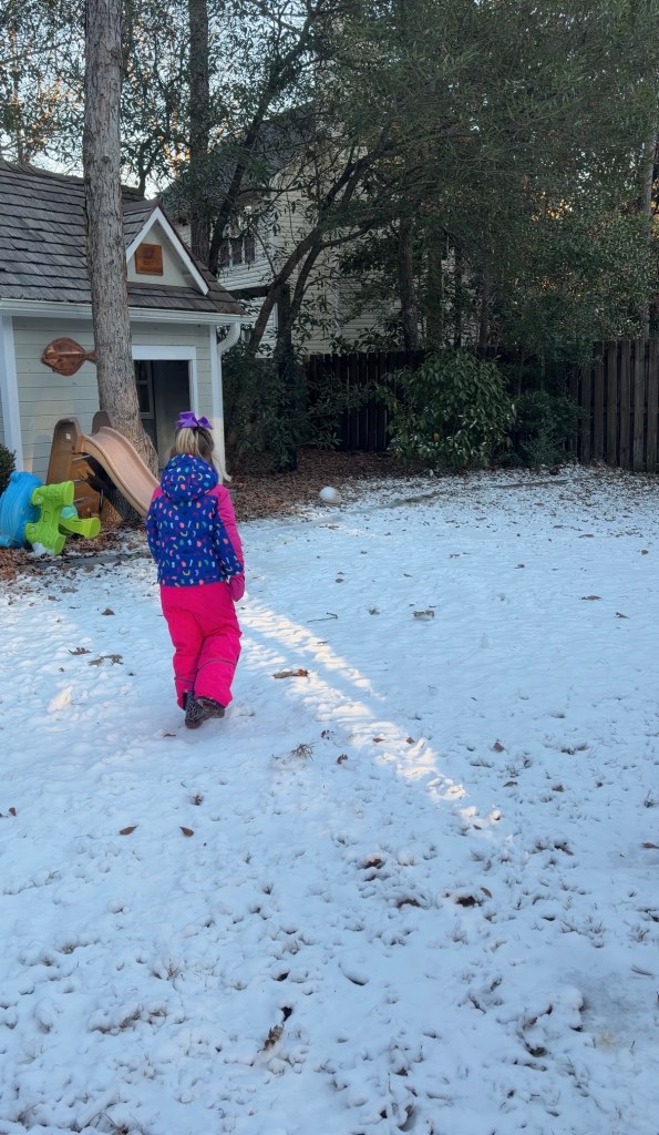A child wearing a colorful jacket and pink pants walks through a backyard covered in snow, with a children's slide visible in the background.