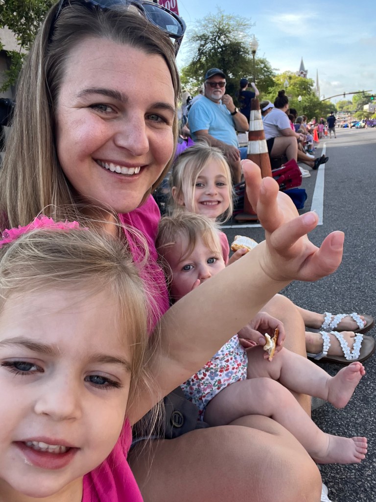 A smiling woman with long hair posing for the camera alongside three young girls, all enjoying a festive outdoor event. The backdrop includes a crowd and trees.