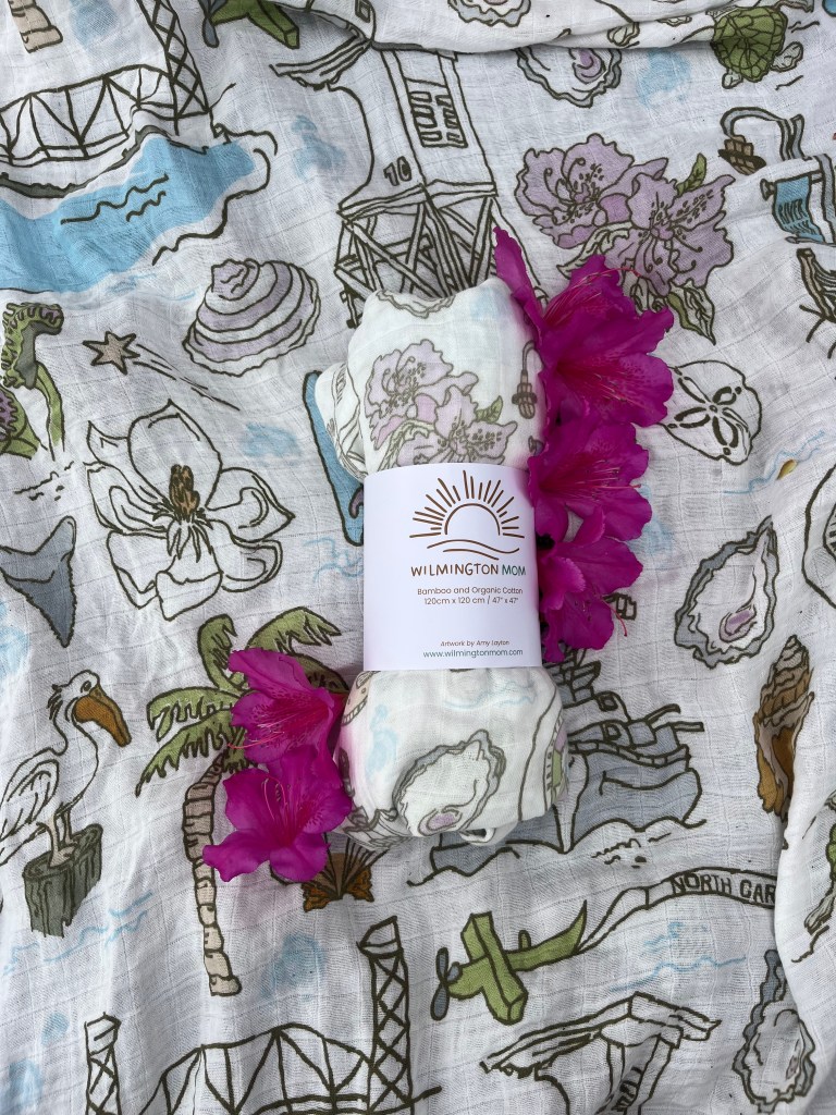 A rolled bamboo and organic cotton blanket with a label reading 'Wilmington Mom' placed on a patterned fabric featuring colorful illustrations of coastal elements, flowers, and palm trees, accompanied by vibrant pink flowers.