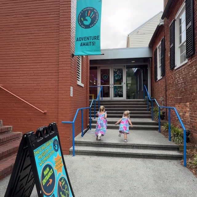 Two children in colorful dresses walking up the stairs towards the entrance of a children's museum, with a sign reading 'Adventure Awaits!' and an additional sign detailing museum information.