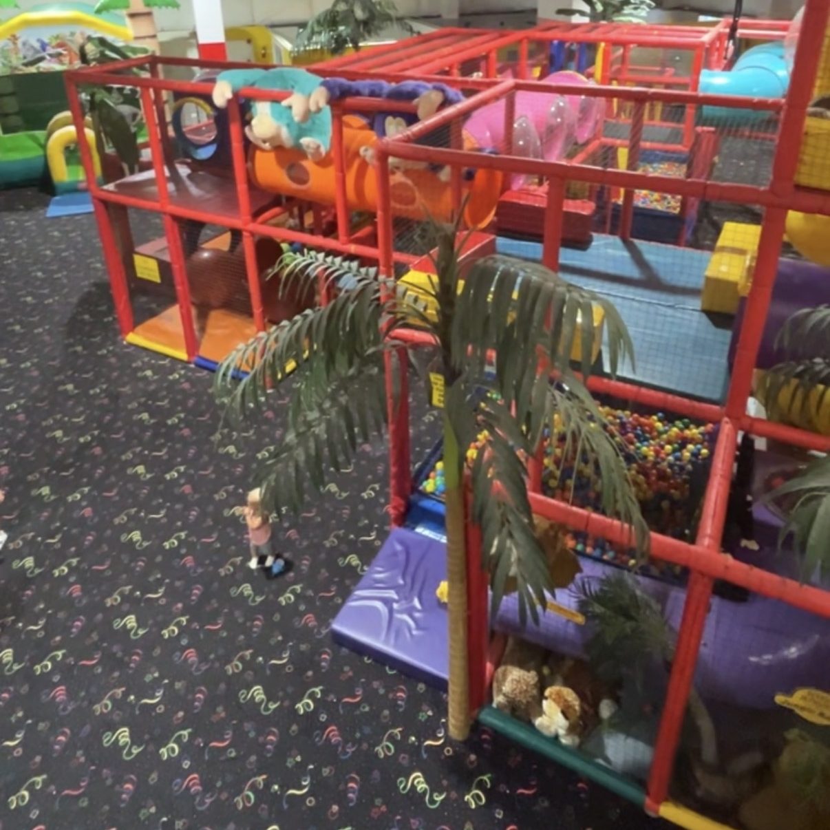An indoor play area featuring colorful climbing structures, slides, and tunnels. There are decorative palm trees and a soft play surface with multicolored patterns. A small child is seen exploring the space.