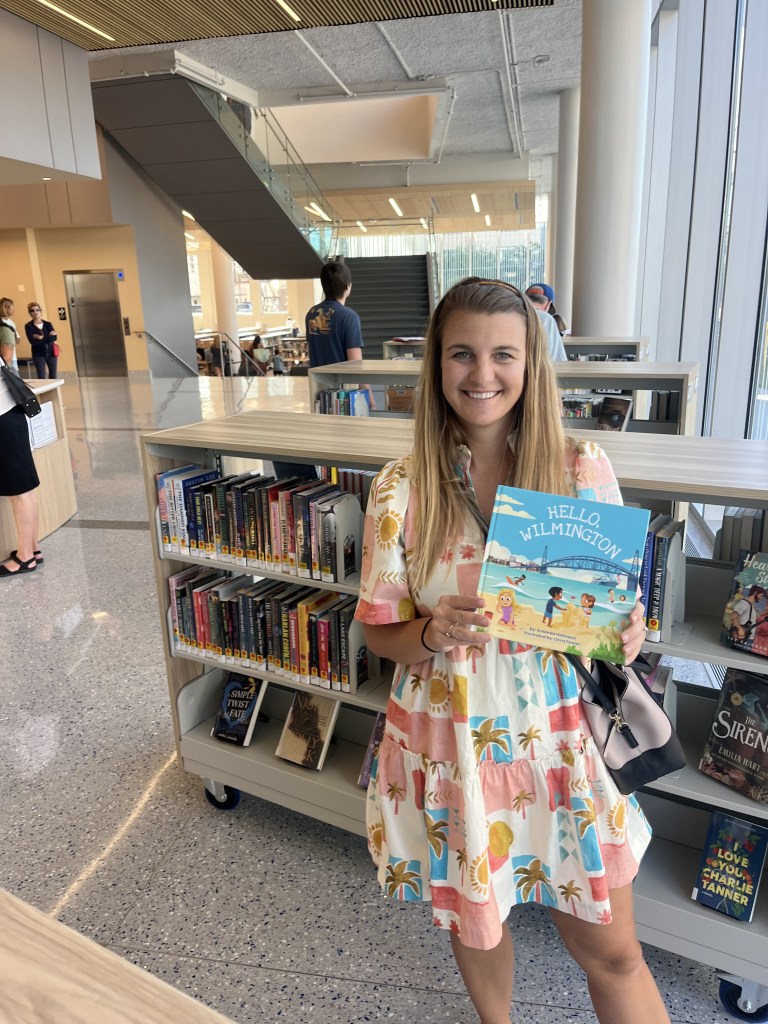 A smiling woman in a colorful summer dress holding a children's book titled 'Hello Wilmington' while standing in a modern library. In the background, there are bookshelves filled with various books and people walking.