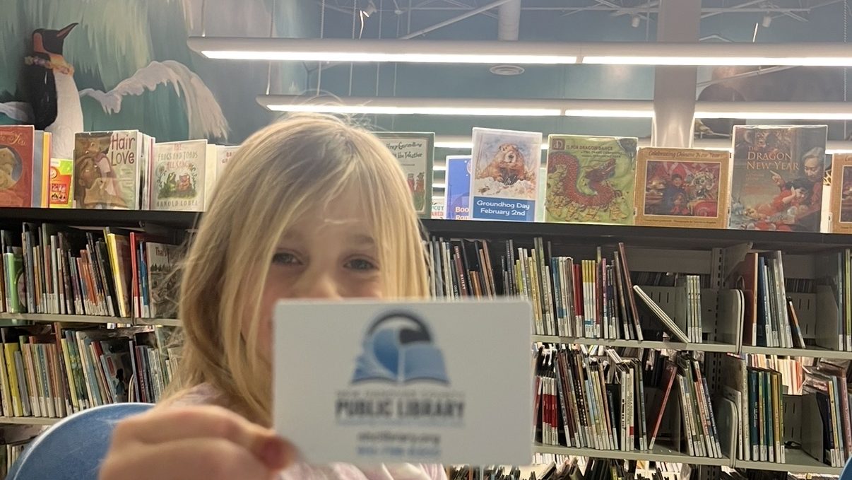 A young child holding a library card in a public library, seated at a table with colorful books nearby.