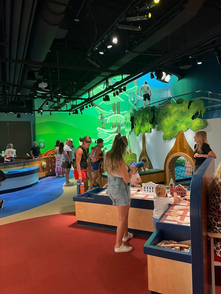 A colorful indoor play area filled with families. Children are engaged with various play stations, including building blocks, while adults supervise and interact. The background features vibrant murals and playful design elements.
