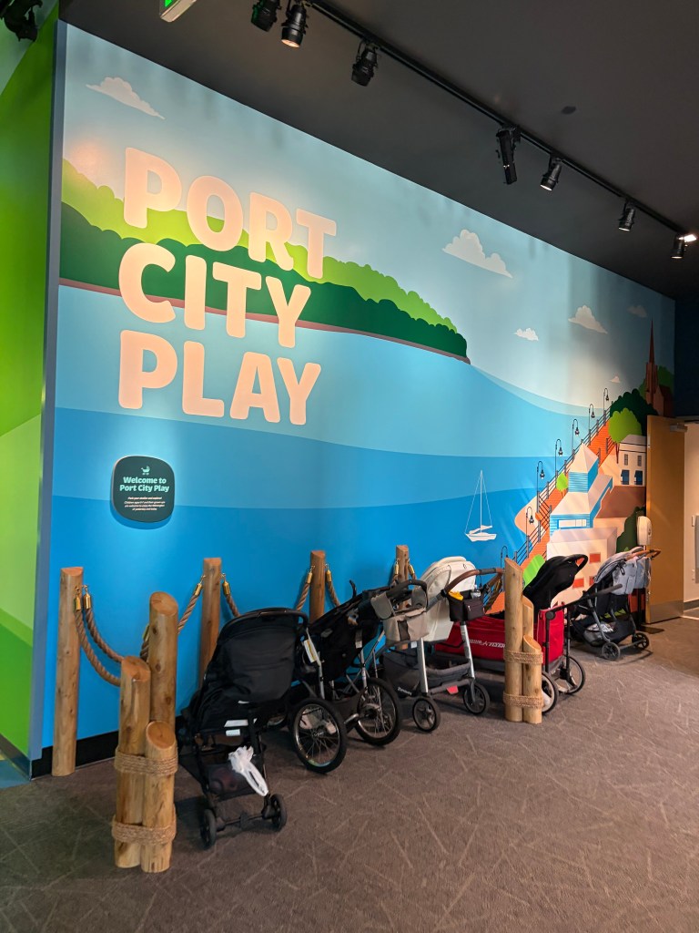 A colorful wall mural featuring the text 'PORT CITY PLAY' with a scenic waterfront illustration, including trees, clouds, and boats. In the foreground, there are several strollers lined up next to wooden posts and ropes.