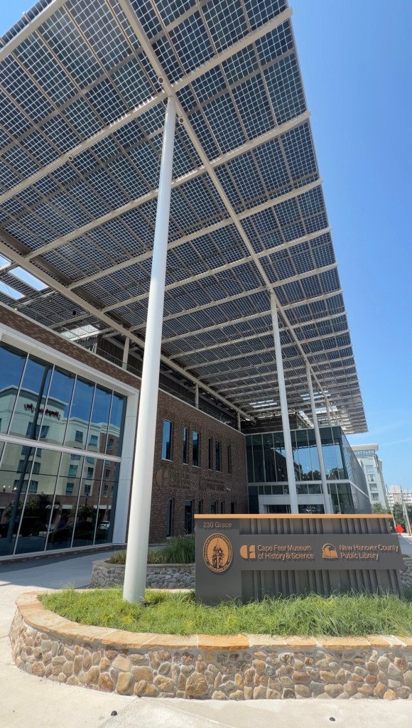 Exterior view of the Cape Fear Museum of History and Science and New Hanover County Public Library buildings, featuring a solar panel canopy and modern architecture.