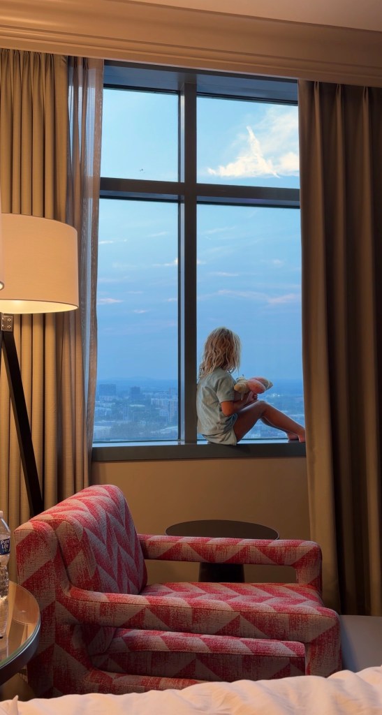 A young child sitting on a windowsill in a hotel room, gazing out at the sky and cityscape, holding a stuffed animal. Soft lighting with curtains and a patterned chair in the foreground.