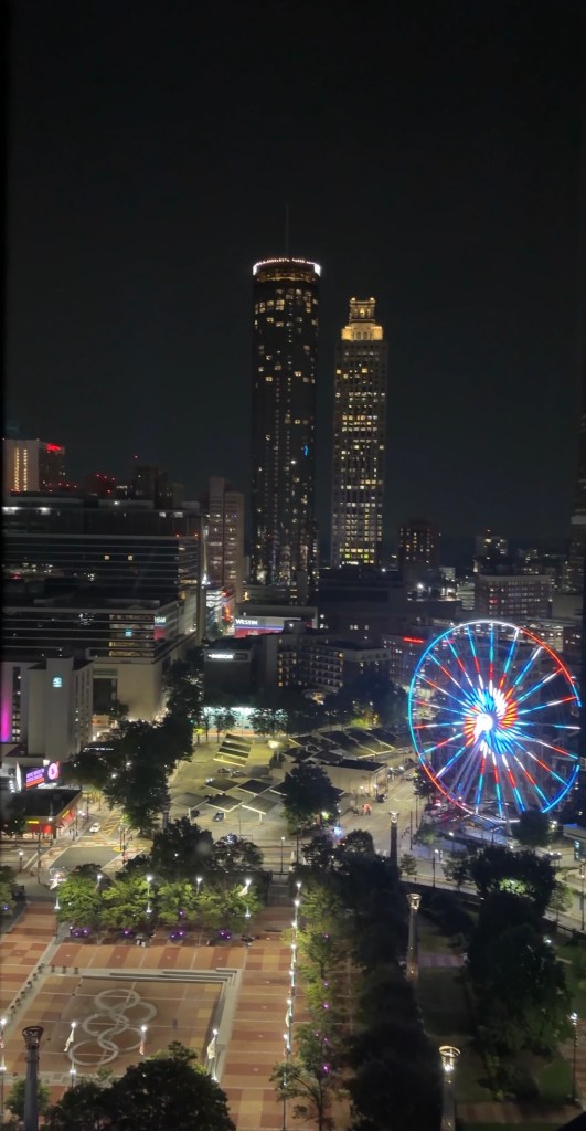 Nighttime view of a city skyline featuring tall illuminated buildings and a colorful Ferris wheel in the foreground.