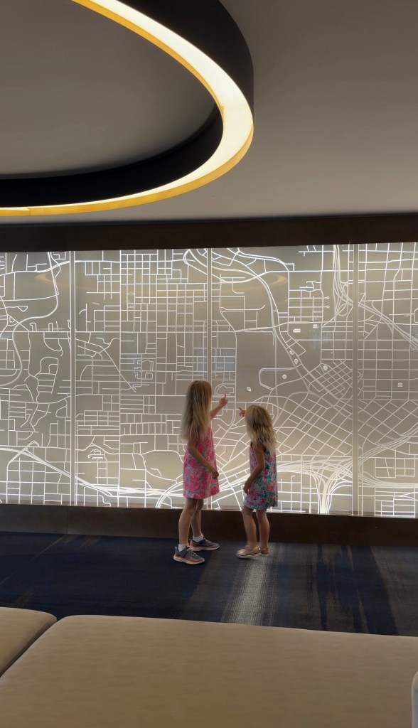 Two young girls interacting with a backlit map display on a wall, indoors, in a modern setting.