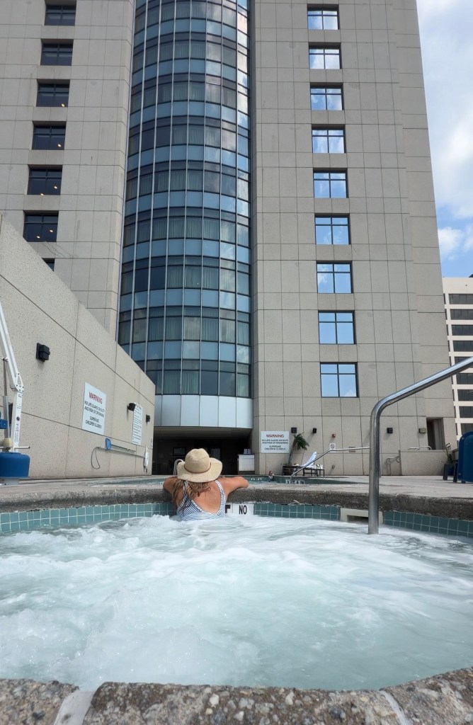 A person with curly hair and a straw hat relaxing in a hot tub with bubbling water, in front of a modern building with large glass windows.