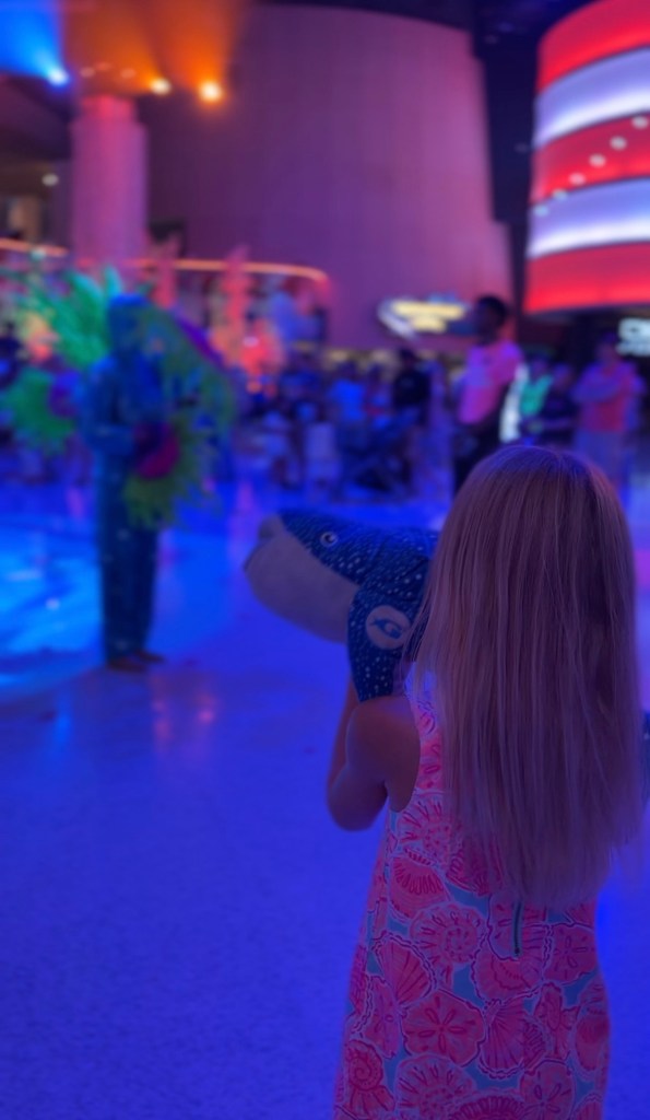 A young child in a colorful dress holds a stuffed shark toy while watching a performance with bright lighting and colorful decorations in the background.