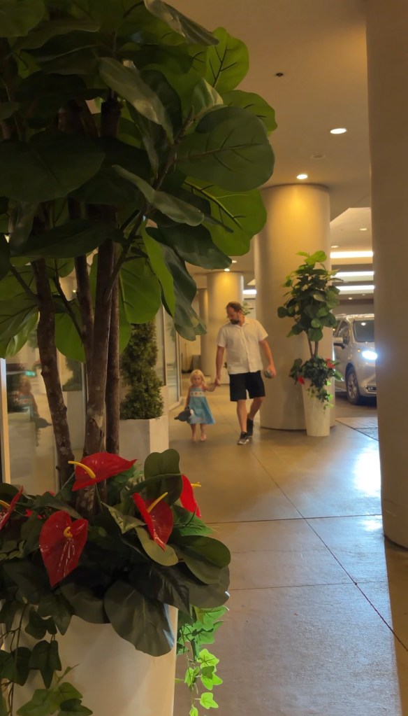 A man and a young girl walking together in a well-lit indoor area, surrounded by potted plants and flowers.